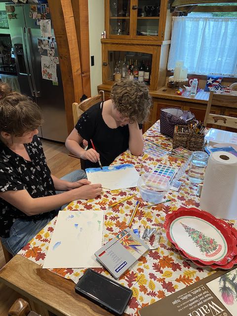 A teen girl and her art teacher sit at a table creating a watercolor painting together. Spread out on the table are various art materials.