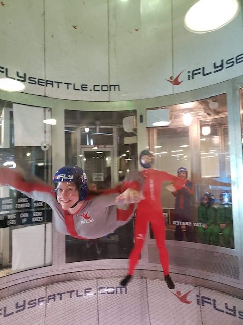 A smiling woman in an indoor skydiving tube, with the staff member in the background.