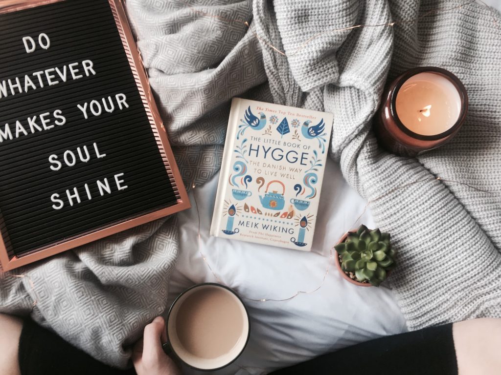 A book about hygge on a blanket, with a candle, a hand holding a cup of coffee, and a sign that says, "Do whatever makes your soul shine."