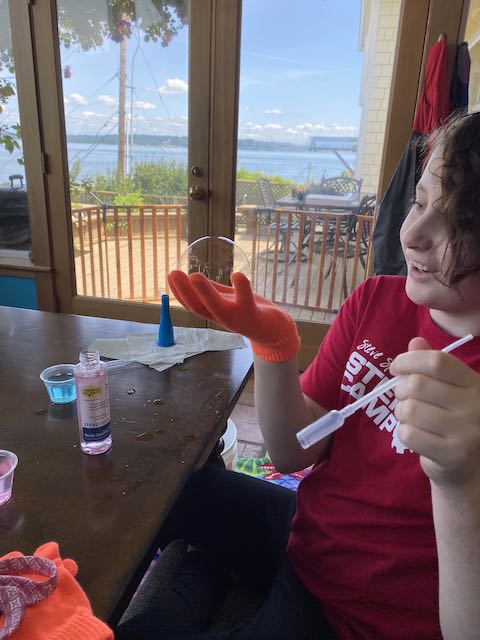An elementary age girl in gloves holding a bubble and smiling. This picture was taken during our STEM camp with Steve Spangler.