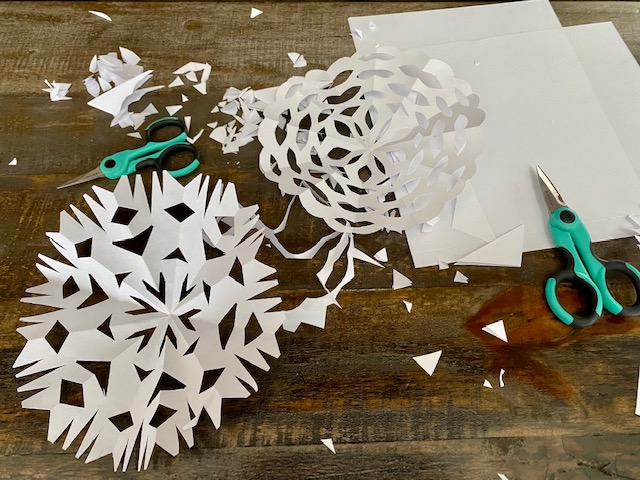 A table with paper, scissors, and two paper snowflakes unfolded with paper trimmings.