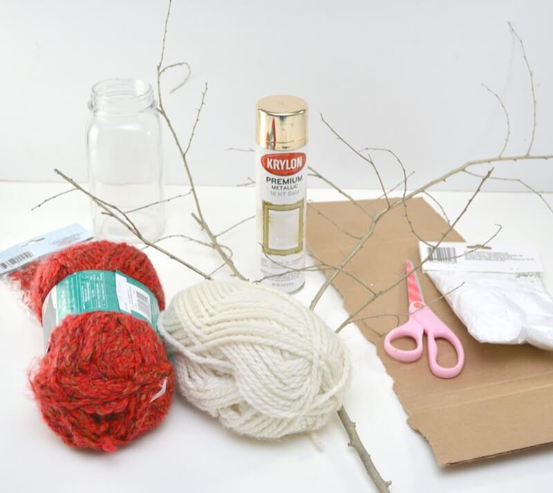 Supplies to make a Valentine tree craft: yarn, branches, scissors, cardboard, paint all laying on a white table.