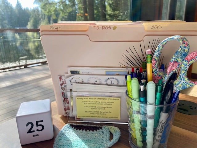 A closeup of the corner of a desk in front of a window. There is a small cube timer, a stand up file holder filled with files, and a cup with pens and pencils.